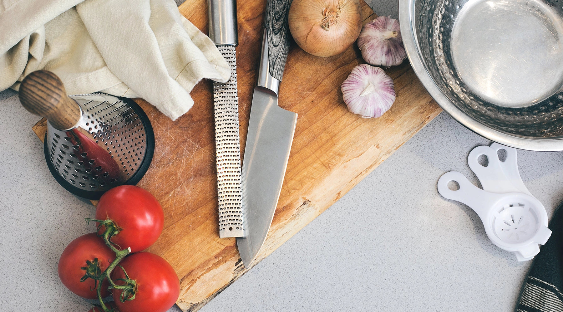 Top-down view of high-quality kitchen knives on a wooden cutting board, displayed alongside a grater, metal mixing bowl, and fresh ingredients like tomatoes, onion, and garlic. Ideal setup showcasing premium chef's knives